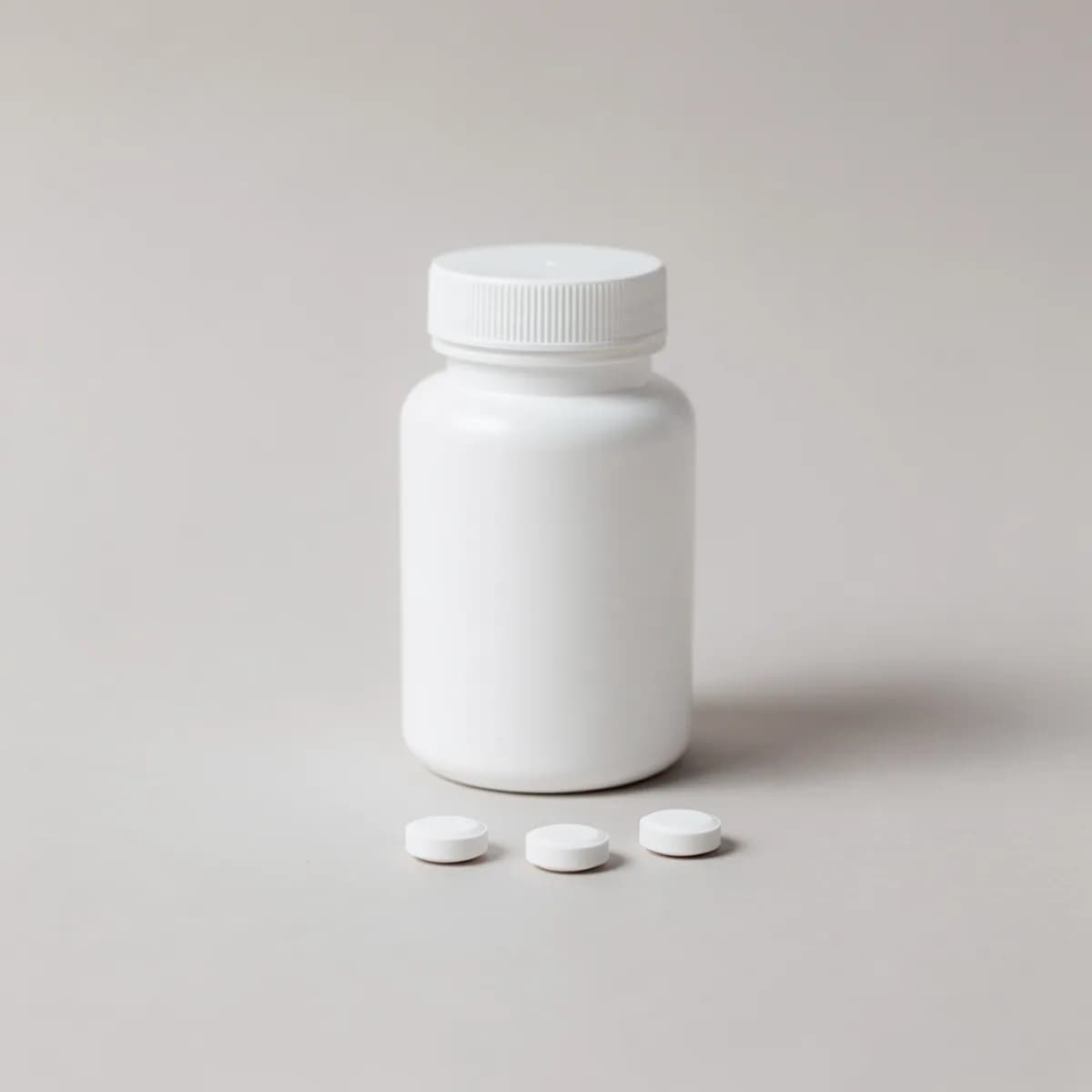 A plain white unlabelled pill bottle with three white tablets beside it on a pale surface.