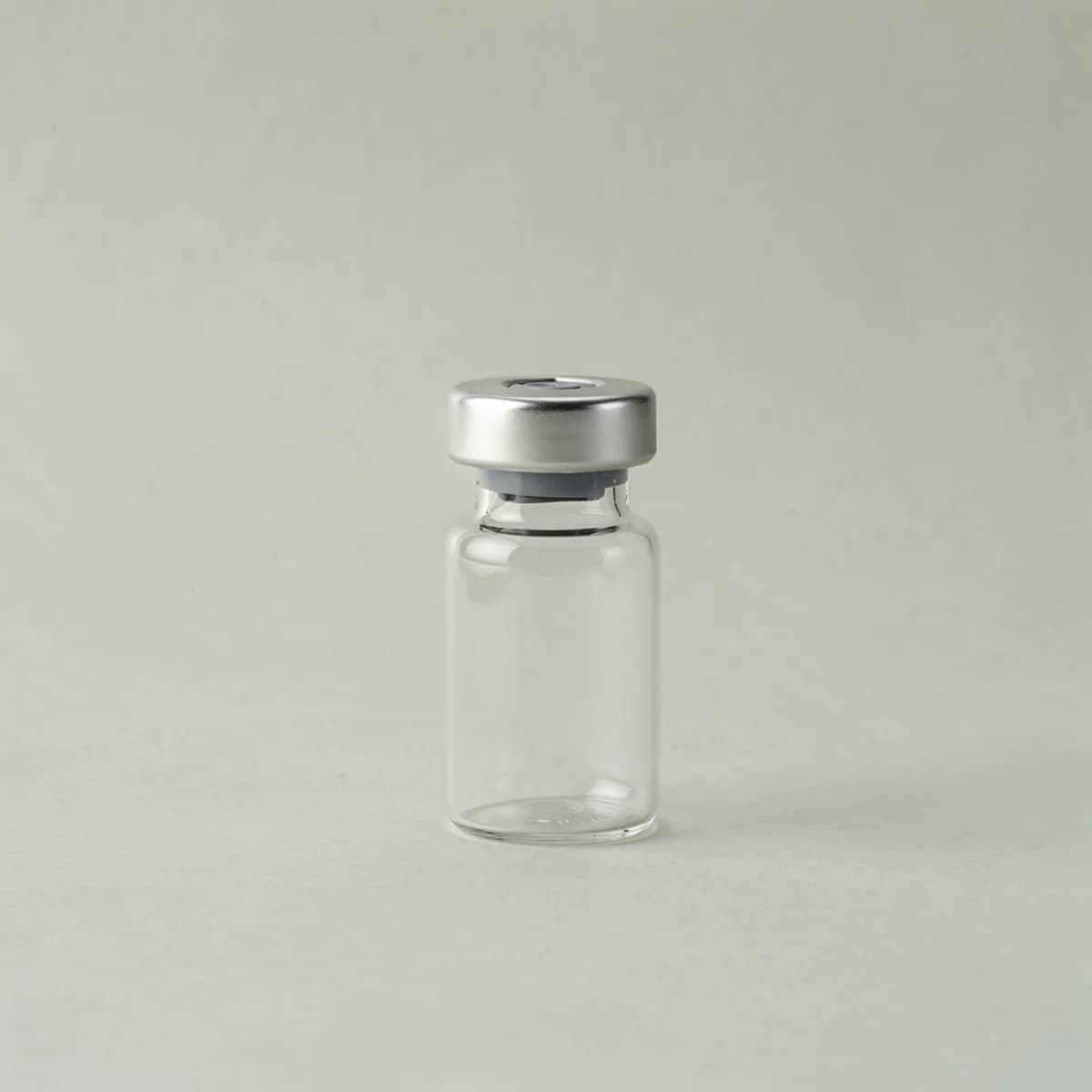 A small clear glass medical vial with a plain silver cap, unlabelled, on a pale surface.
