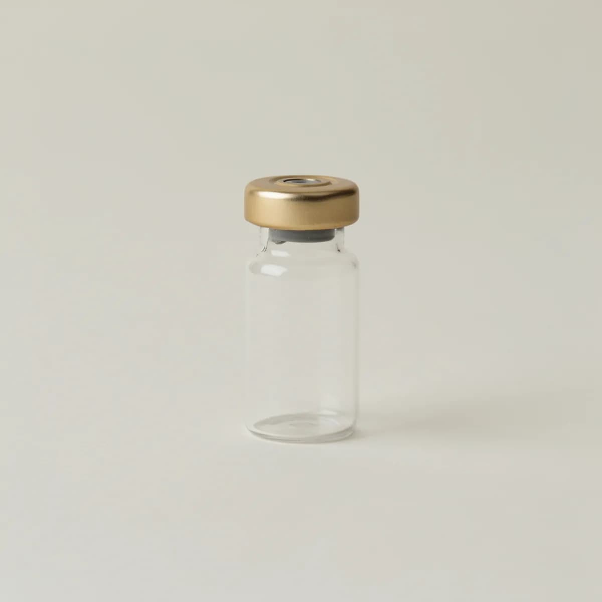 A small clear glass medical vial with a plain gold cap, unlabelled, on a pale surface.