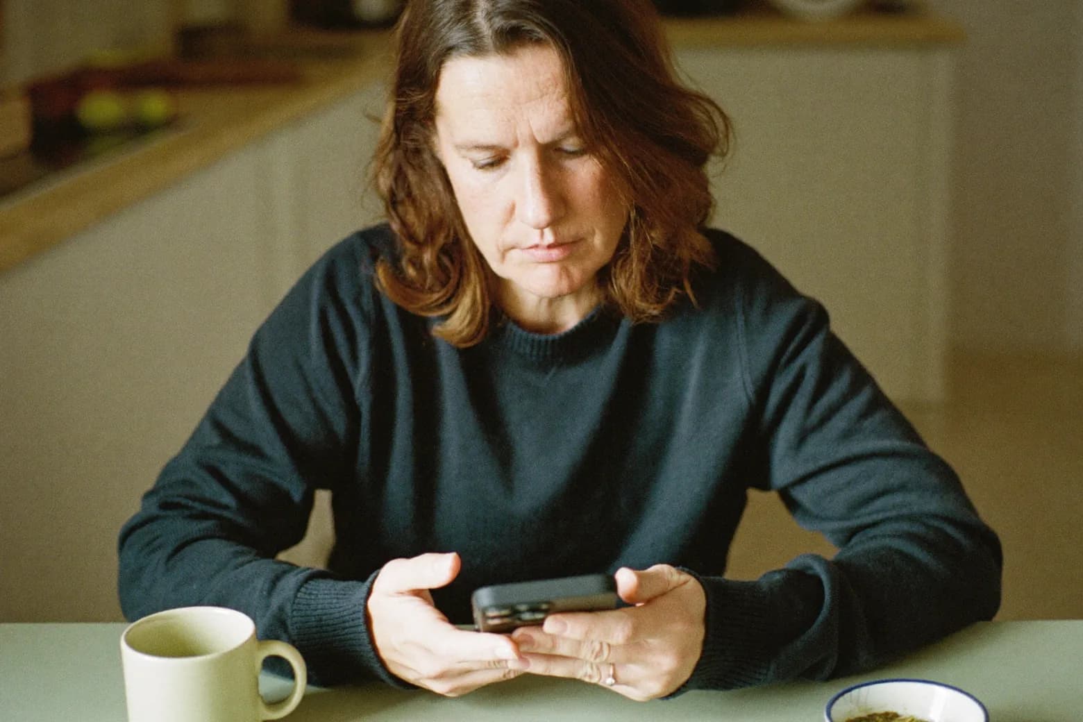 A woman sits at her kitchen table using her phone.