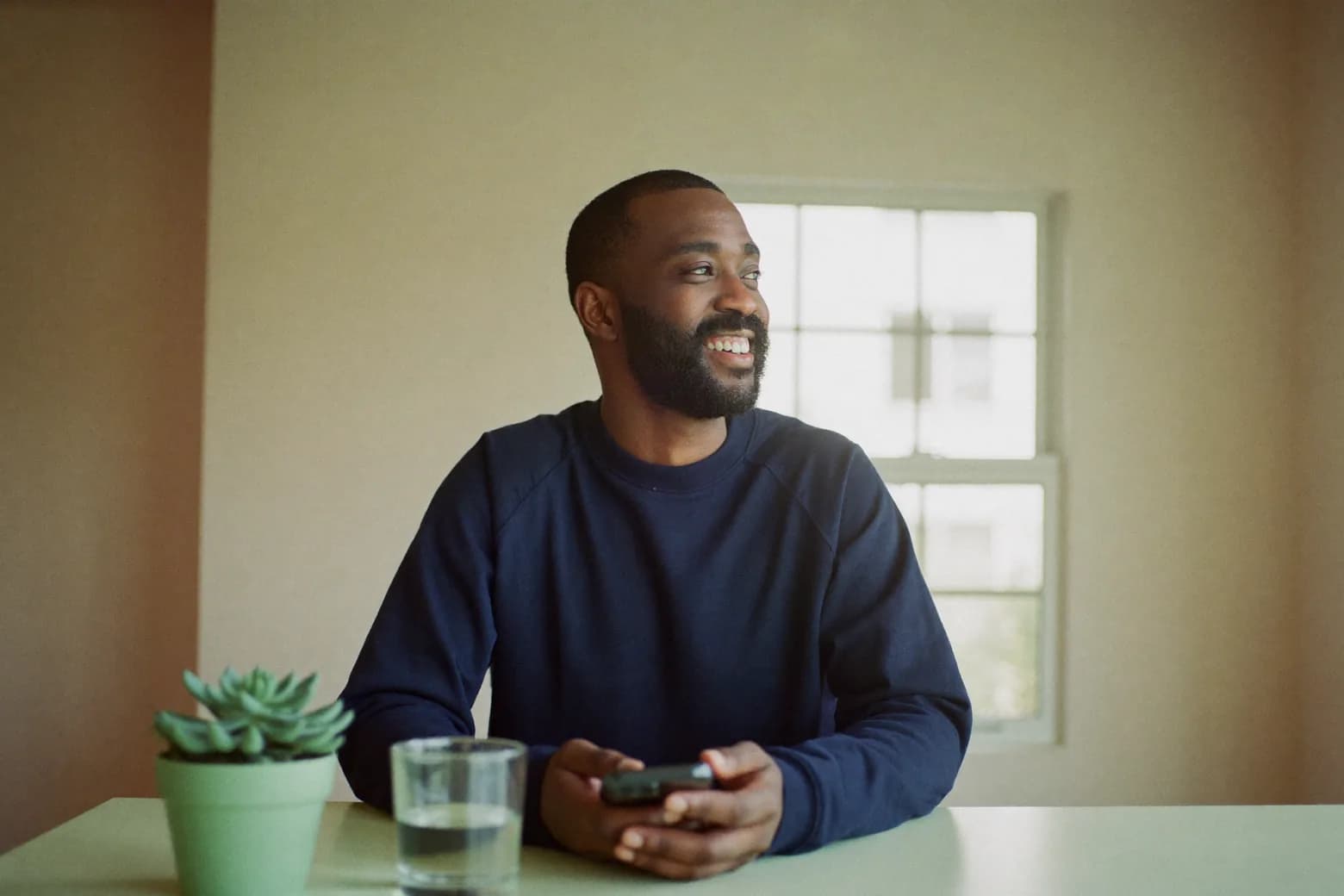 A man sits at his kitchen table holding his phone, smiling as he looks toward a window.